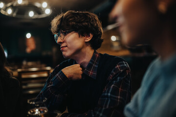 A young man wearing a plaid shirt and glasses sits in a cozy, nighttime restaurant setting with warm lighting.