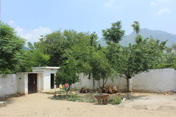 A rural compound featuring a whitewashed house, a dirt courtyard, trees, and a low wall, set against a backdrop of green mountains under a bright sky