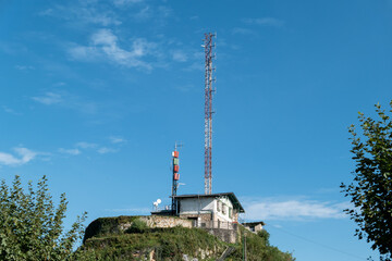 Obraz premium Imponente torre de telecomunicações sobre uma estrutura metálica, erguida sobre um pequeno edifício de controlo situado no alto de uma colina coberta de vegetação, sob um céu azul