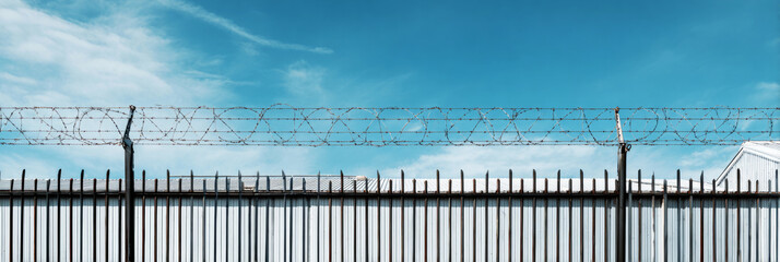A high steel fence with barbed wire encloses a warehouse. The photo shows a clear blue sky above and distant buildings in the background during the day, banner