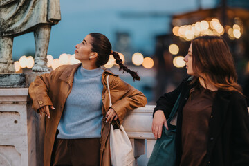 Two youthful friends in an urban setting lean on a marble railing, chatting as warm city lights glow in the background. A casual, candid moment of friendship and city life.