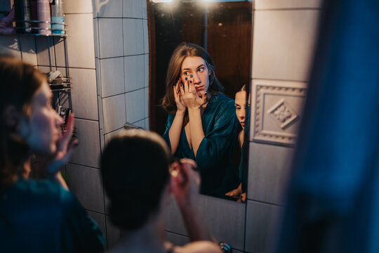 A young woman in a teal robe applies makeup in a tiled bathroom, seen through a mirror as a friend looks on. The scene conveys self-care, preparation, and intimate everyday beauty. - Powered by Adobe