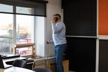 Small business owner of a blinds retail shop standing by a window with a roller shade, talking on...