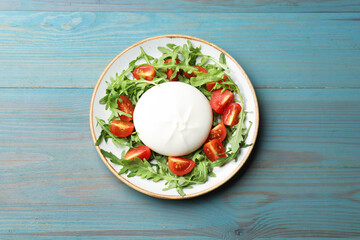 Delicious burrata cheese with tomatoes and arugula on light blue wooden table, top view