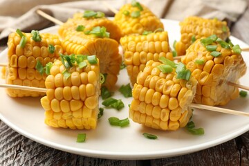 Skewers with pieces of boiled corncobs and green onion on wooden table, closeup