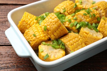 Pieces of boiled corncobs with green onion in baking dish on wooden table, closeup