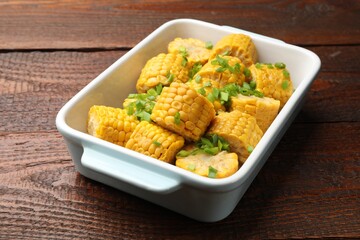 Pieces of boiled corncobs with green onion in baking dish on wooden table, closeup