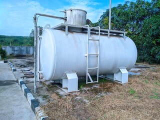 Horizontal, white industrial fuel storage tank with ladder, piping, and small upper vessel, sitting on concrete supports outdoors.
