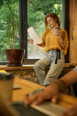 Red-haired woman reading a paper by a large window