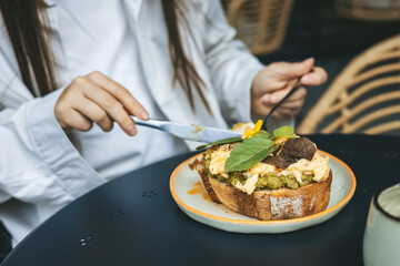 Close-up shot of a person wearing a white shirt cutting scrambled eggs on avocado toast with a knife and fork during brunch