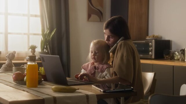 Medium shot of busy working mother using laptop and speaking by phone while sitting at desk at home with cute little daughter in lap, copy space