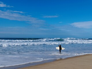 Woman in a wetsuit with a surfboard under her arm stands in front of the storm-tossed Atlantic Ocean in Mimizan Plage, France.