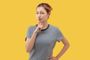 Playful young Asian woman making silence gesture with finger on lips on yellow background.