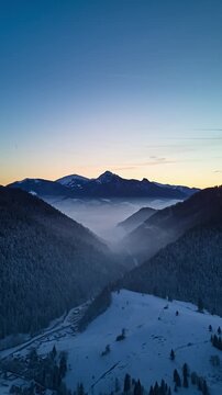 Aerial evening hyperlapse flying forward over snowy forest valley with mist and distant mountain ridge
