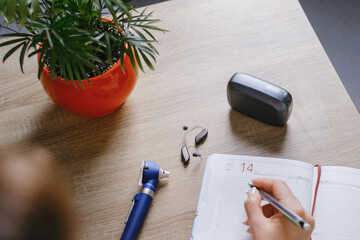 Modern digital hearing aids, otoscope, and open planner on wooden table in audiology clinic....