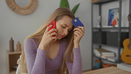 Woman indoors holding phone and credit card in living room wearing purple shirt with concerned expression next to guitar and books on shelf at home.