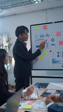 Confident Asian manager leading a business meeting, pointing at charts on a whiteboard. Team of colleagues brainstorming new marketing strategies