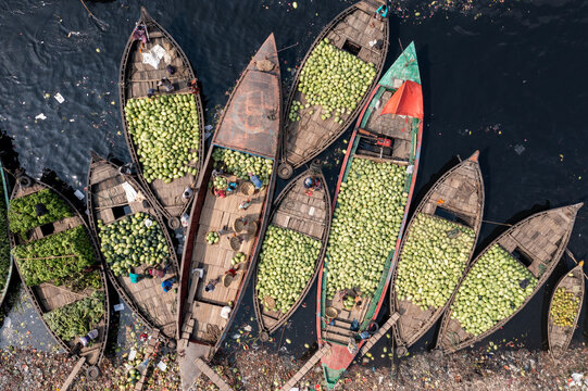 Aerial view of boats brimming with vibrant green produce contrast against the dark waters and trash-strewn banks, Dhaka, Dhaka Division, Bangladesh.