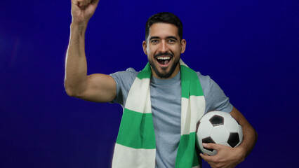 Excited man holding soccer ball wearing green scarf over blue background celebrating victory fan...