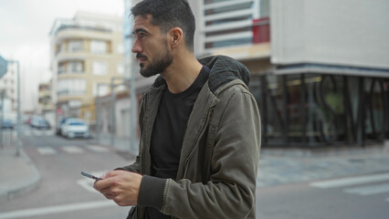 Young man using smartphone on urban street, wearing a jacket and carrying backpack, surrounded by...