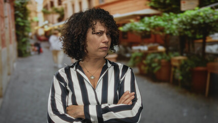 Hispanic woman in black and white striped blouse crosses arms on cobblestone street lined with...