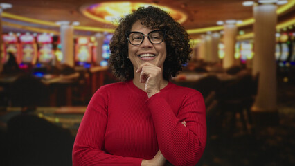 Woman stands with hand on chin leaning at casino slot machine row under chandelier lighting;...