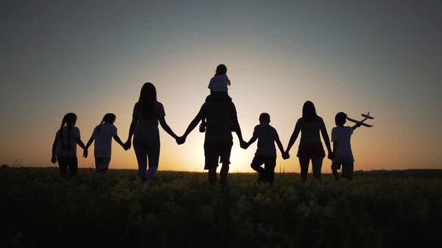 Family walking hand in hand across field at sunset, silhouette of parent and child holding toy plane, group together enjoying outdoor play and unity with horizon glow and tall grass under warm light