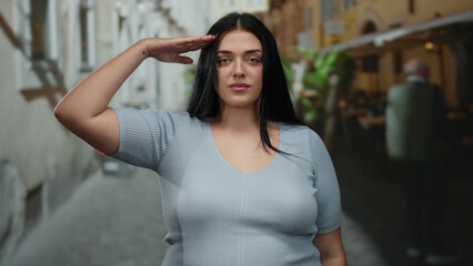 Woman saluting on city street gives confident expression while standing outdoors showcasing youthful style and curvy figure in urban environment.