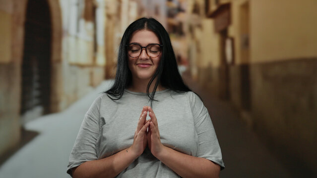 Woman smiling on a quiet city street wearing glasses and a grey shirt on a bright day with a blurred background showcasing charming urban architecture.