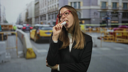 Fototapeta premium Woman holding wrapped snack to mouth on busy city street, arms crossed and thinking; pensive urban pause.