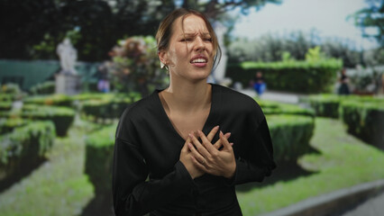 Fototapeta premium Woman clutching chest for heartattack in forest pathway beside hedge with hands pressed to sternum and grimace; medical emergency pain.