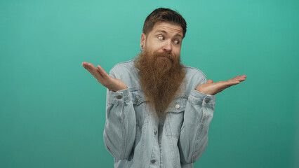Bearded young caucasian man in denim jacket shrugs shoulders palms up in studio with teal backdrop;...
