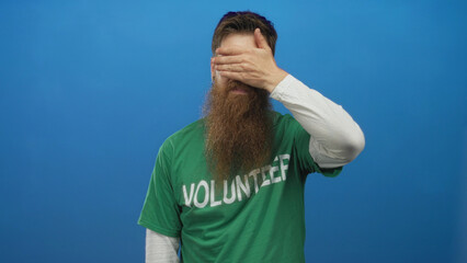 Naklejka premium Man in green volunteer shirt covers eyes with hand against blue studio wall; uncertainty reflection.