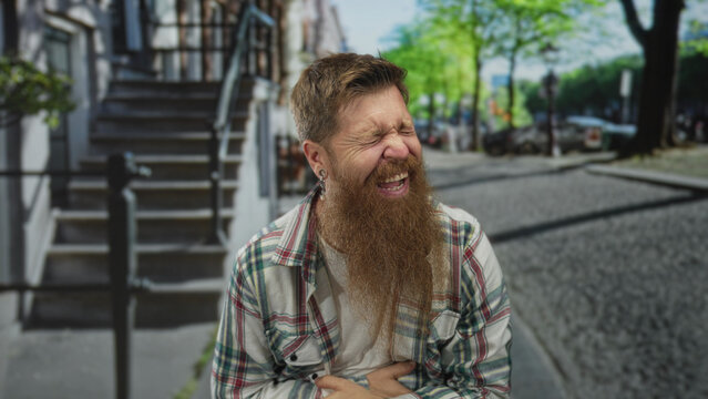 Man with long beard and plaid shirt pointing finger toward camera in urban street with building facade visible; amusement.