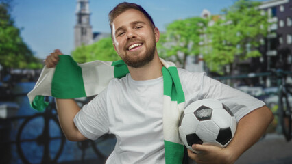 Naklejka premium Man smiling while holding soccer ball and waving green scarf on street with hand and forearm visible by canal railings; celebration support.
