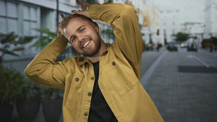 Man face and hands making bunny ears gesture while smiling broadly, wearing yellow jacket and black shirt on a city street lined with planters; playful joy.