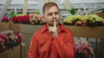 Man in orange prisoner jumpsuit pressing finger to lips for silence amid vibrant flower market display in building; silence secrecy restraint.