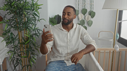 Man sitting holding smartphone beside baby crib inside a building room looking at screen; fatherhood bonding affection.