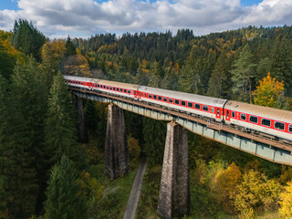 Aerial view of a vibrant red and white train traversing a towering stone viaduct amidst a dense forest in the Cierna voda valley, Cierna voda, Zilinsky kraj, Slovakia.