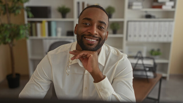 Man holding eyeglasses and resting chin on hand at office desk, smiling while looking at monitor; calm confidence.