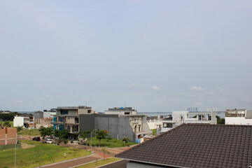 Fototapeta premium Brown tiled roof of a house in the foreground with a view of a residential area and other buildings under construction, with the sea on the horizon.