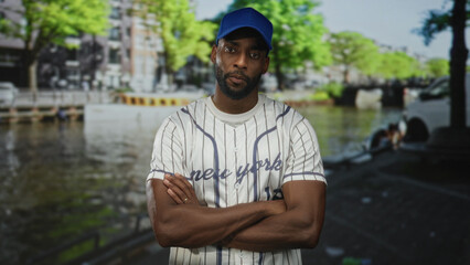 Fototapeta premium Man in baseball jersey with arms crossed standing by an amsterdam canal on street; urban confidence.