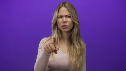 Fototapeta premium Blonde caucasian woman points finger at camera in purple studio with serious expression; accusation anger.