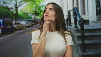 Woman touches chin on street under green canopy near parked cars and building steps; contemplation.