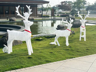 Three decorative white reindeer figures with tall antlers and red collars adorned with golden bells stand on a lush green lawn next to a tranquil pond.