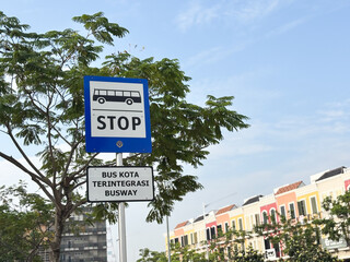 A bright, outdoor, eye-level shot capturing a blue and white bus stop sign set against a clear blue sky and lush green foliage.