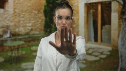 Young hispanic woman scientist in white lab coat raises palm stop gesture at stone building...