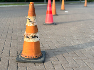 A close-up, ground-level shot of a weathered orange traffic cone with a black base, standing on a paved area made of interlocking bricks.