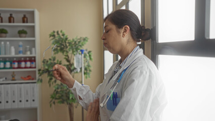 Woman doctor in white coat holding stethoscope pinching nose by clinic window in building corridor;...