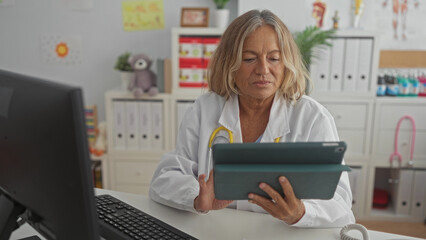 Senior woman pediatrician holds tablet and taps screen while typing at computer in pediatric...